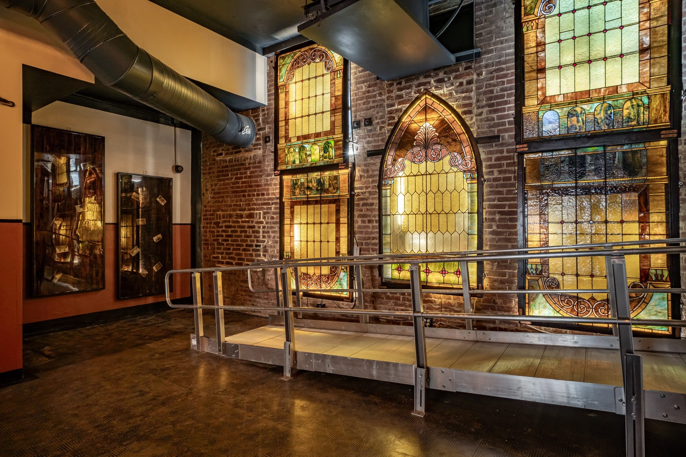 Main hall at Journeyman's Hall with a wall of antique stained-glass windows above a raised stage, exposed brick and warm lighting