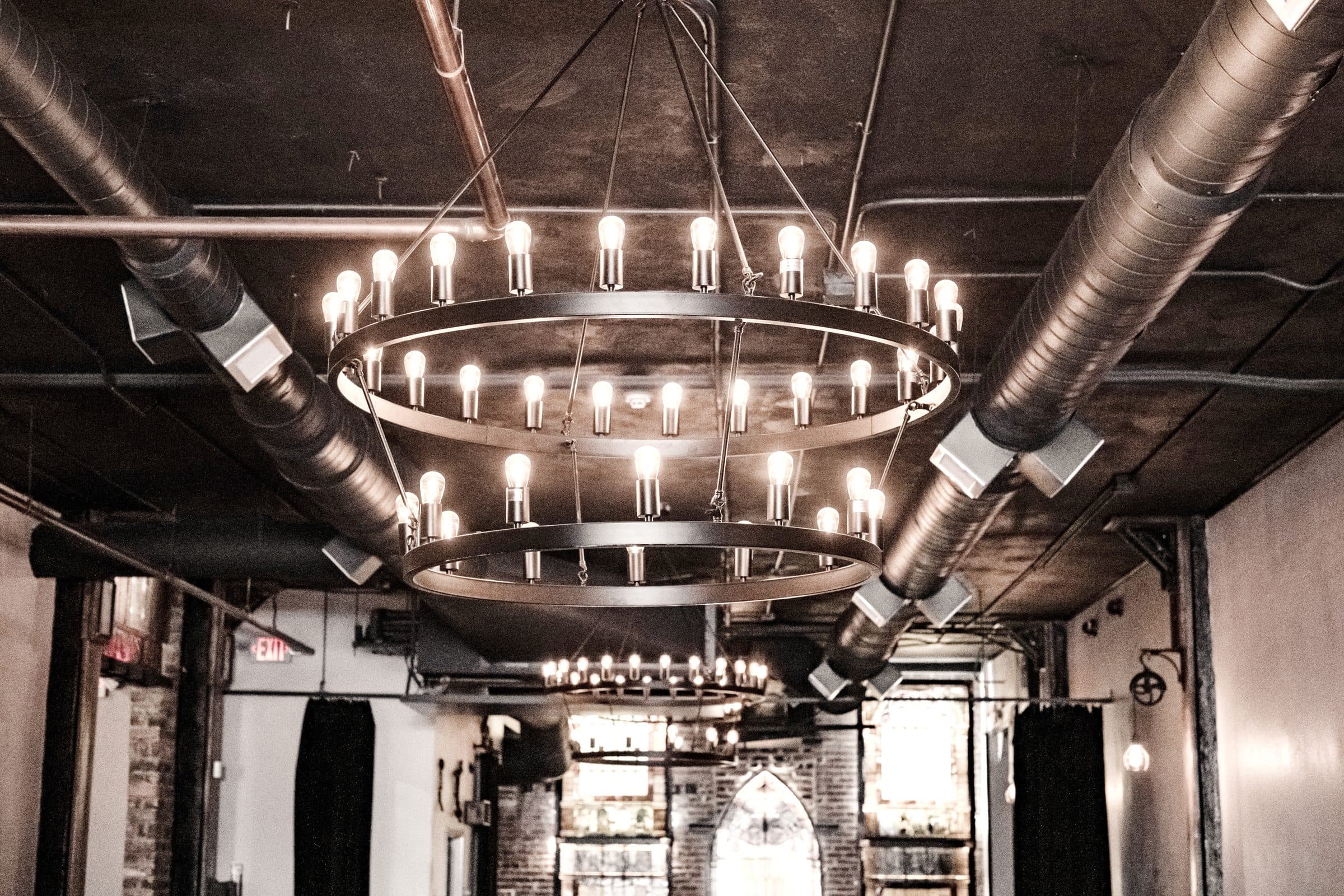 Iron ring chandelier with exposed bulbs below industrial ductwork